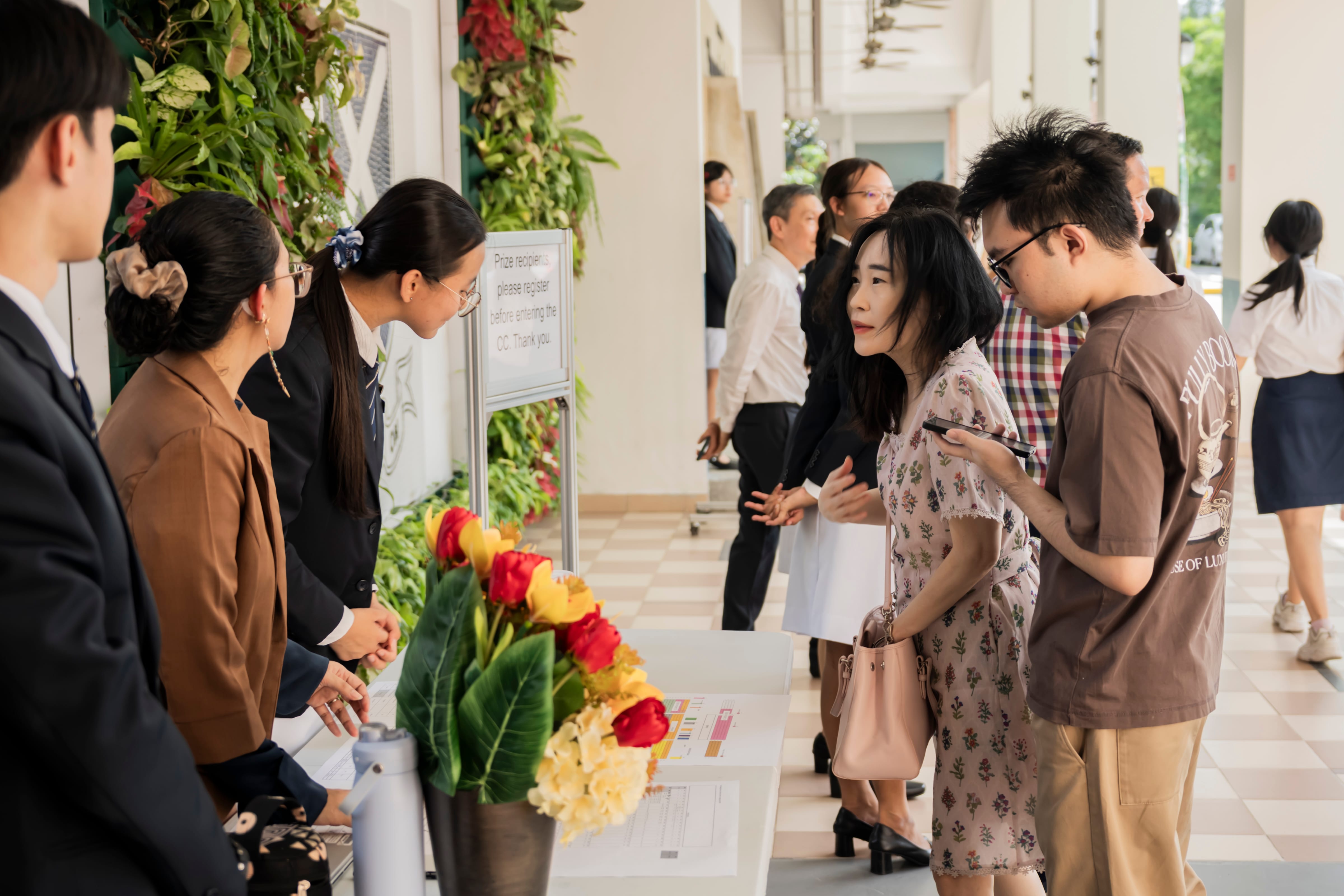 Guests check in at a registration counter decorated with flowers before an event along an outdoor school corridor.